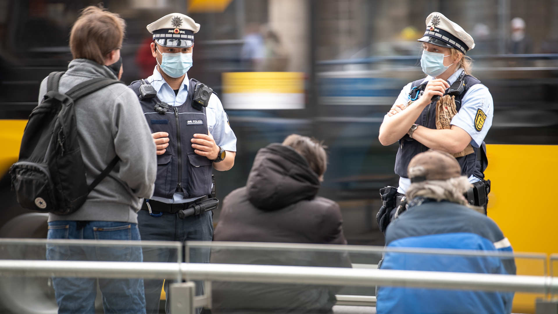 Zwei Polizeibeamte mit Gesichtsmaske sprechen an einer Haltestelle drei Männer an. | dpa