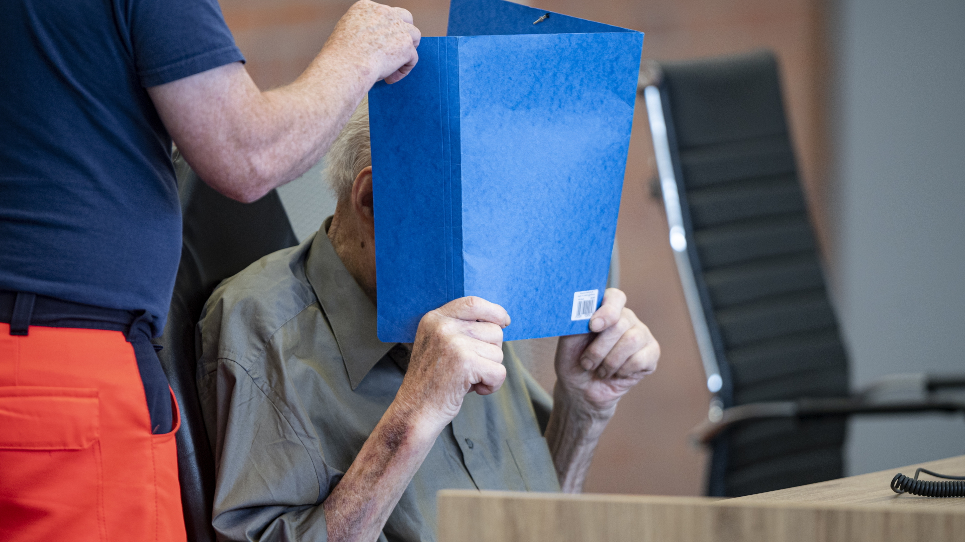 Der 101 Jahre alte angeklagte frühere Wachmann im KZ Sachsenhausen hält sich vor dem Gericht in Neuruppin einen blauen Schnellhefter vor das Gesicht. | dpa