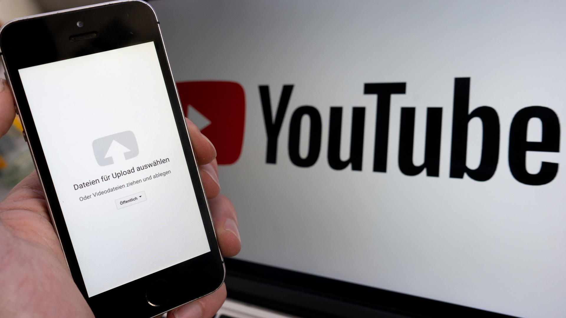 Ein Upload-Fenster der Video-Plattform Youtube ist auf einem Smartphone vor einem YouTube-Logo auf einem Bildschirm zu sehen. | dpa